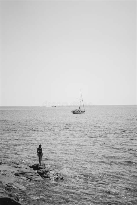 Female Wearing Bikini Standing On A Rock By The Body Of Water With A Sailboat Floating In The