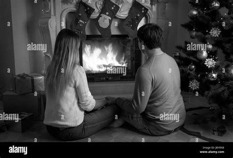 Black And White Image Of Young Couple Sitting Under Christmas Tree And Looking At Burning