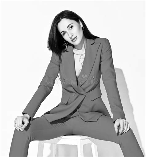 Black And White Portrait Of Young Brunette Woman In Official Pantsuit Sitting On A Stool With