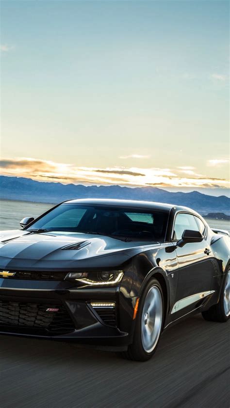 A Black Chevrolet Camaro Driving Down The Road At Sunset With Mountains