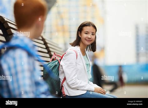 Asian Schoolgirl With Backpack Sitting On The Bench And Talking To Her Classmate After School