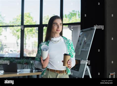 Brunette Queer Person Holding Blueprint And Coffee To Go In Office