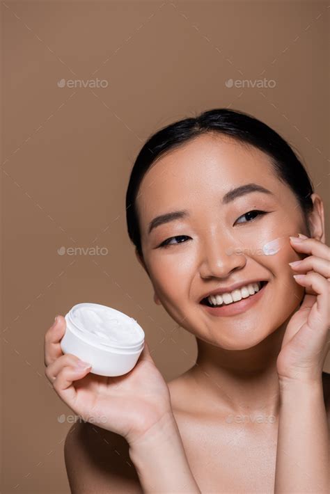 Positive Asian Woman With Naked Shoulders Applying Face Cream Isolated