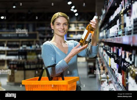 Junge schöne blonde Frau wählt Wein in einem Supermarkt In der Alkoholabteilung stehend mit