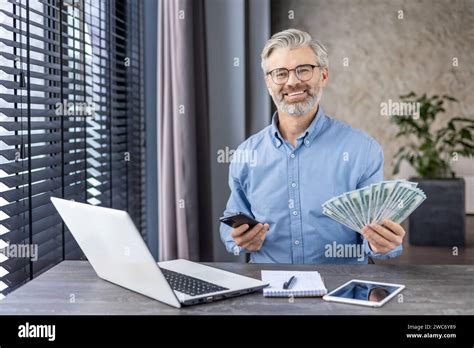 Smiling Mature Businessman With Phone And Cash At Desk Conveying