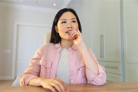 Thoughtful Asian Woman Sitting At Table At Home Making A Video Call