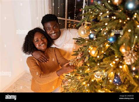 Smiling Millennial Black Wife And Husband Decorate Christmas Tree With Lights And Toys In Cozy