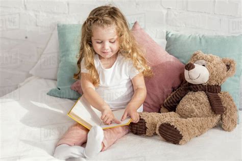 Adorable Cheerful Kid Playing With Teddy Bear And Reading Book On Bed