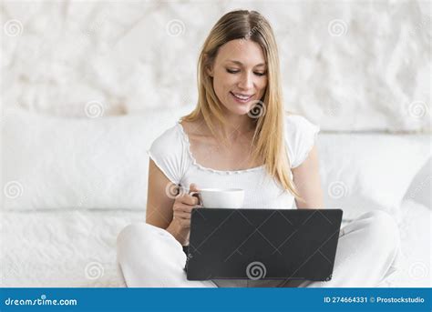 Cheerful Busy Millennial Caucasian Blonde Lady Sitting On Bed With Coffee Cup And Watch Video On