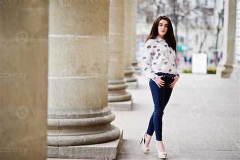 Young stylish brunette girl on shirt, pants and high heels shoes, posed near stone columns ...