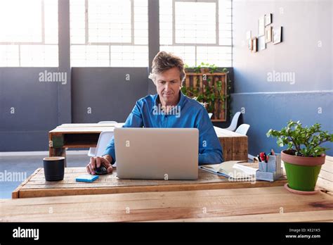 Smiling Mature Businessman Sitting Alone At His Desk In A Large Modern Office Working Online
