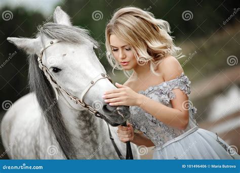 La Belle Fille Blonde Dans La Robe Frotte Un Cheval Gris Sur La Nature Dedans Photo Stock