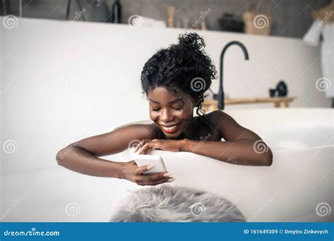 Smiling Dark Skinned Woman Lying In Bath And Using Smartphone Stock