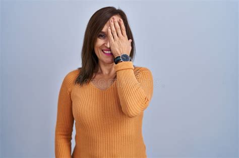 Middle Age Brunette Woman Standing Wearing Orange Sweater Covering One Eye With Hand Confident