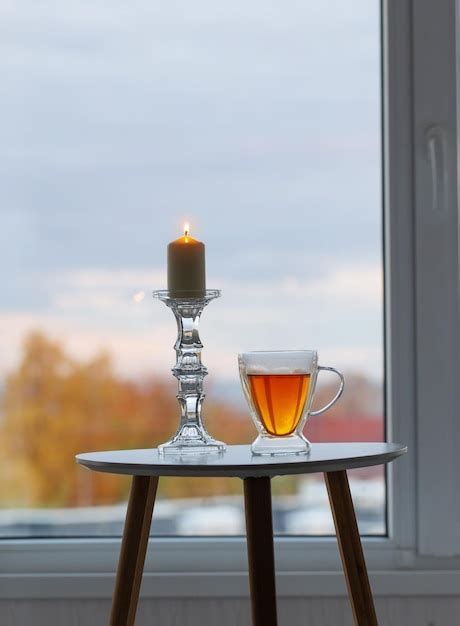 Taza termo de vidrio con té y velas encendidas en la ventana de fondo