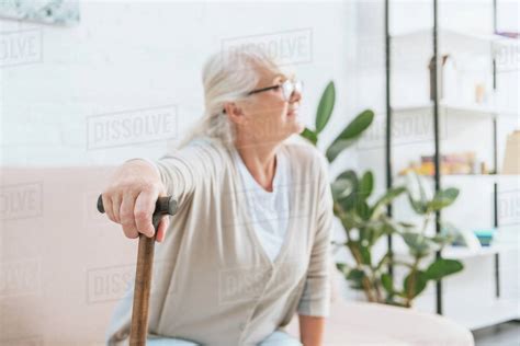 Senior Woman In Eyeglasses Holding Walking Stick And Looking Away While