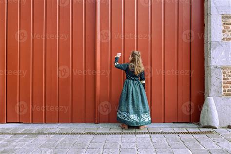 A Little Girl In Medieval Dress Knocking On The Big Red Gate Of The Old