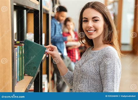 Young Smiling Woman Student Standing In Library Holding Book Stock