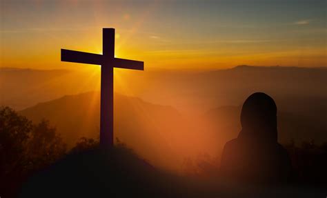 Silhouette Of Woman Holding A Bible Praying In Front Of The Cross On Sunset Background