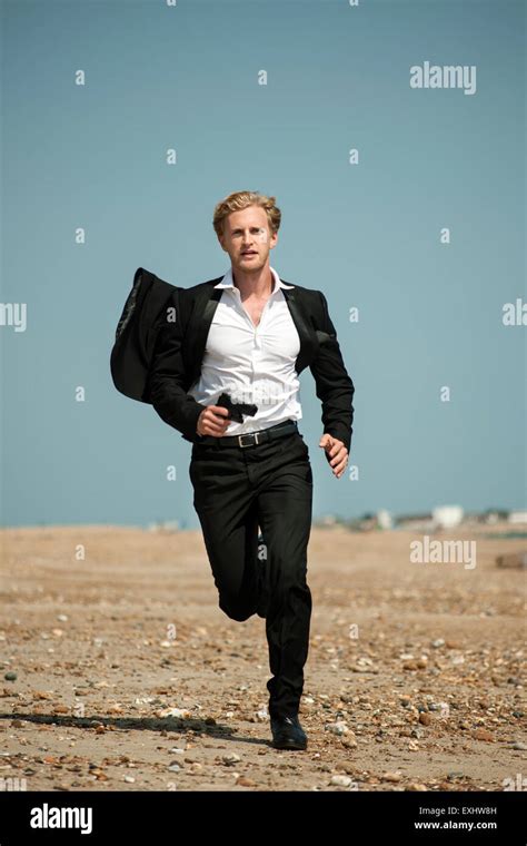 Man In A Black Suit Running After Someone On The Beach Holding A Gun