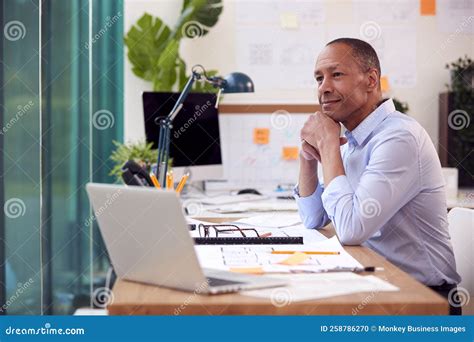 Mature Male Architect Working In Office At Desk Working On Plans For