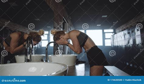 Beautiful Tall Sporty Blonde Woman Washing Face Looking In The Mirror In Dark Gym Locker Room