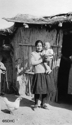 An Old Black And White Photo Of A Woman Holding A Baby In Front Of A Hut