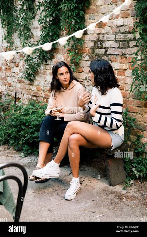 Female Friends Talking To Each Other While Sitting On Bench Near Brick