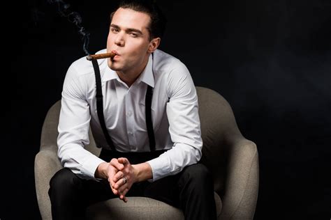 Handsome man smoking cigar while sitting in armchair with clenched hands isolated on black Free ...