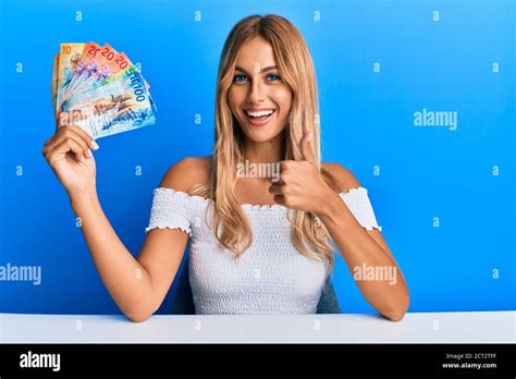 Schöne blonde junge Frau hält schweizer Franken Banknoten lächeln glücklich und positiv Daumen