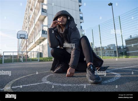 Portrait Of Woman In Overcoat With Hood Sitting On Basketball Court