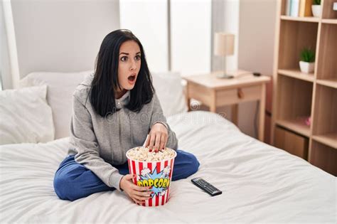 Hispanic Woman Eating Popcorn Watching A Movie On The Bed In Shock Face Looking Skeptical And