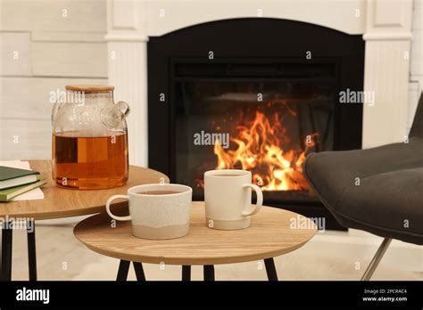 Cups Of Hot Drink And Teapot On Wooden Tables Near Decorative Fireplace