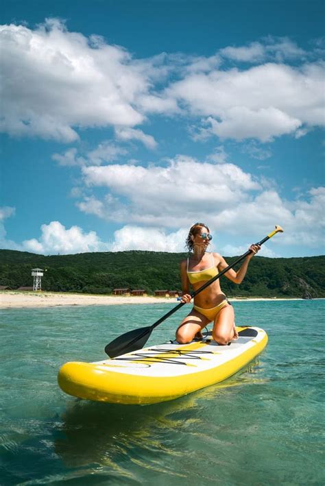 Tanned Woman In Bikini Sitting On Surfboard With Paddle Relaxing At Sea