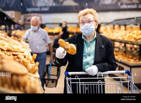 Older Woman With Glasses Chooses Buns And Bread In Supermarket Bakery