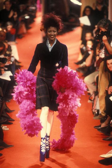 A Woman Walking Down A Runway In A Skirt And Jacket With Pink Feathers