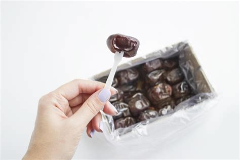 Female Hand Holds Dried Date Palm Fruits Above Box On White Background