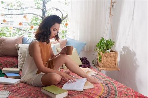 Jeune Femme Lisant Un Livre Assis Sur Son Lit Avec Une Tasse De Boisson