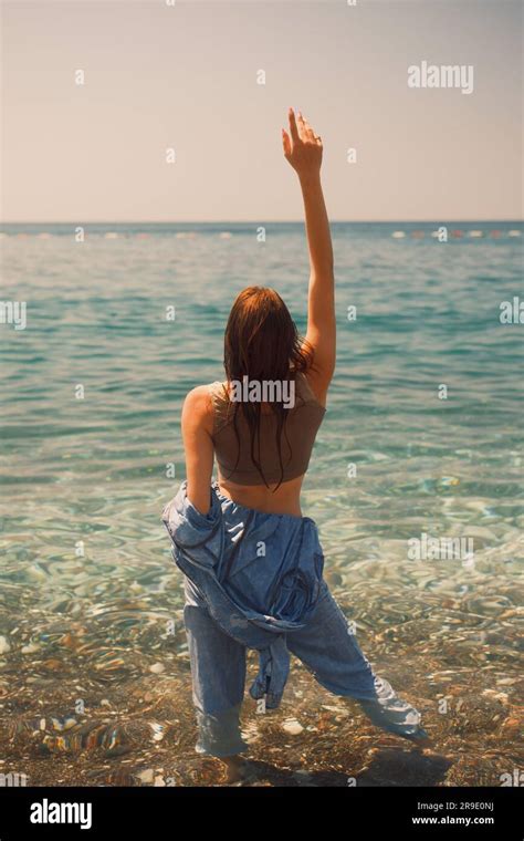 A Girl In Wet Denim Overalls Stands With Her Back To The Camera Her