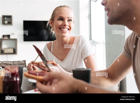 Happy Blonde Woman Looking At Her Man While They Eating Breakfast Together In Kitchen Stock