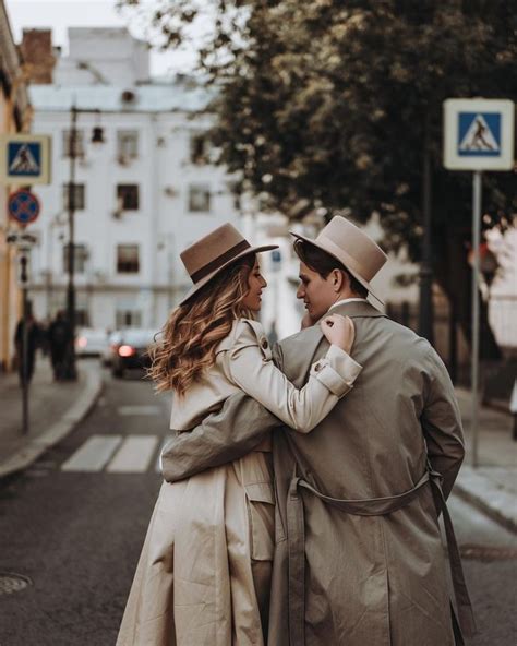 A Man And Woman In Trenchcoats Are Standing On The Side Walk With Their