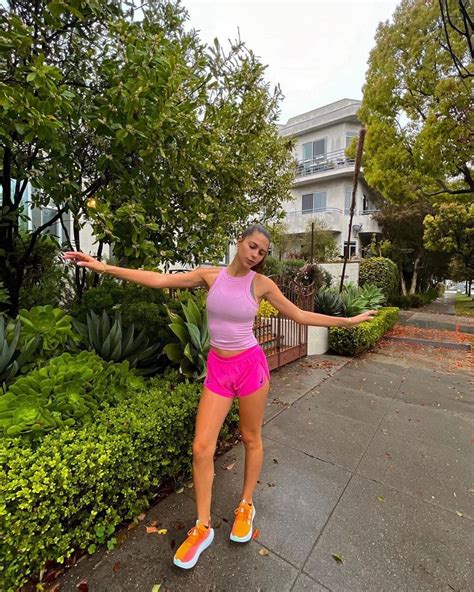 A Woman In Pink Shirt And Shorts Standing On Sidewalk Next To Bushes