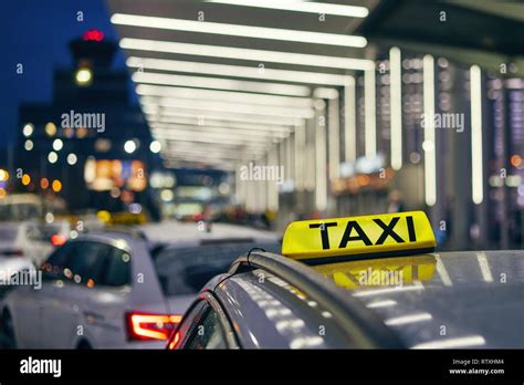 Lighting Taxi Sign On The Roof Of Car Against Airport Terminal At Night
