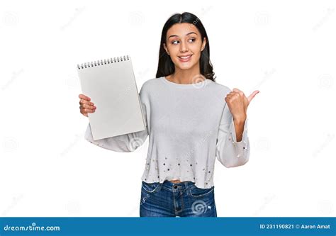 Young Hispanic Girl Holding Notebook Pointing Thumb Up To The Side