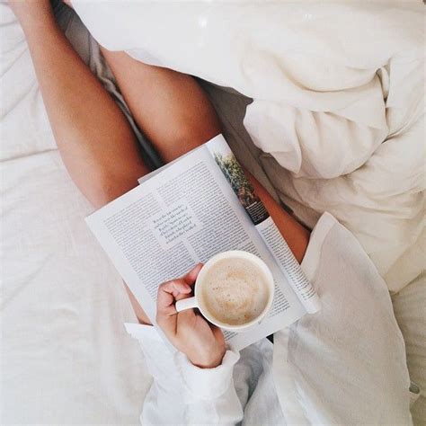 A Woman Laying In Bed Reading A Book And Holding A Cup Of Coffee With