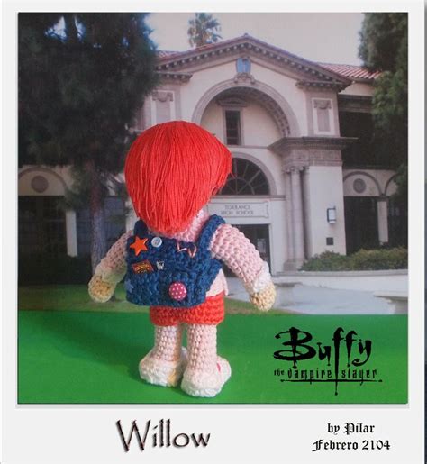 A Crocheted Doll With Red Hair And Blue Overalls Stands In Front Of A House