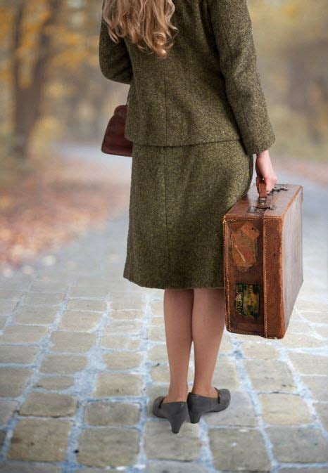 A Woman In A Green Coat And Hat Holding A Brown Suitcase While Talking