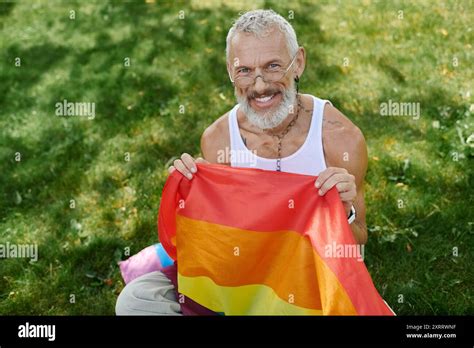 A Mature Gay Man With A Grey Beard And Tattoos Smiles As He Holds A