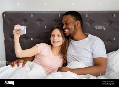 Millennial Multiracial Couple With Smartphone Taking Selfie In Bed