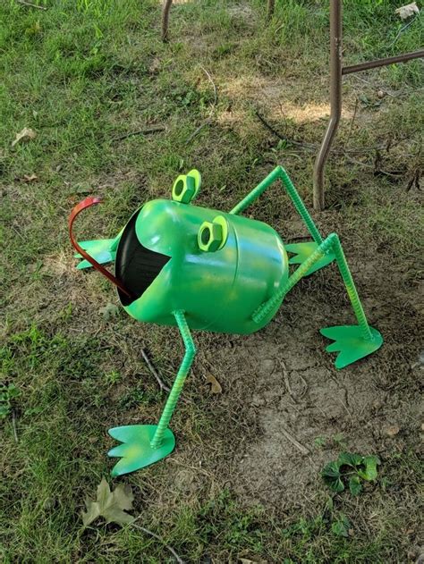 A Green Plastic Frog Sitting On Top Of A Grass Covered Field Next To A Tree